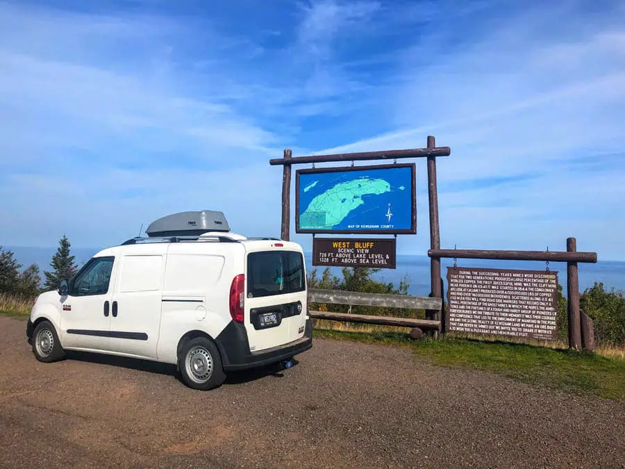 Image of a campervan at top of Brockway Mountain in front of a sign that reads "West Bluff Scenic View" 