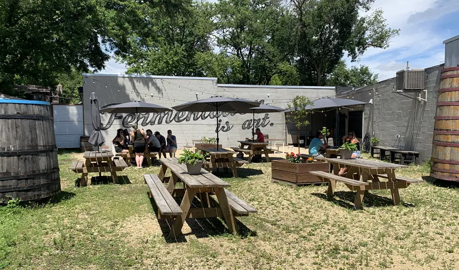 Image of outdoor patio at Fuck Factory Geuzeria in Madison WI. Image is of picnic tables with black umbrellas and a wall in the background with the words "fermentation is art"