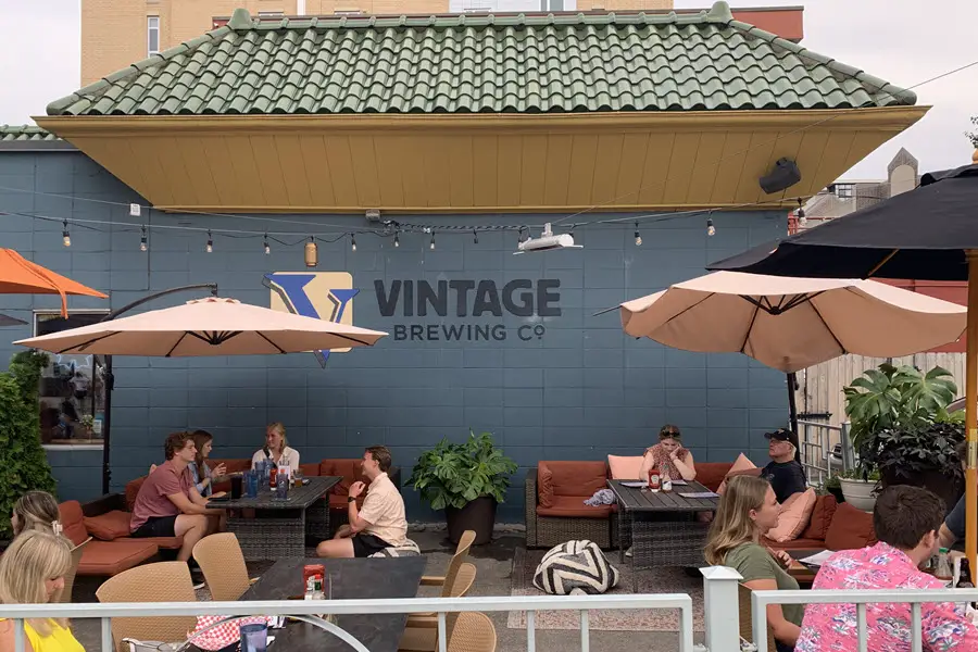 Image of patio at Vintage Brewery in Madison WI. People sitting at tables with a blue wall in the background that reads "Vintage Brewing Co"