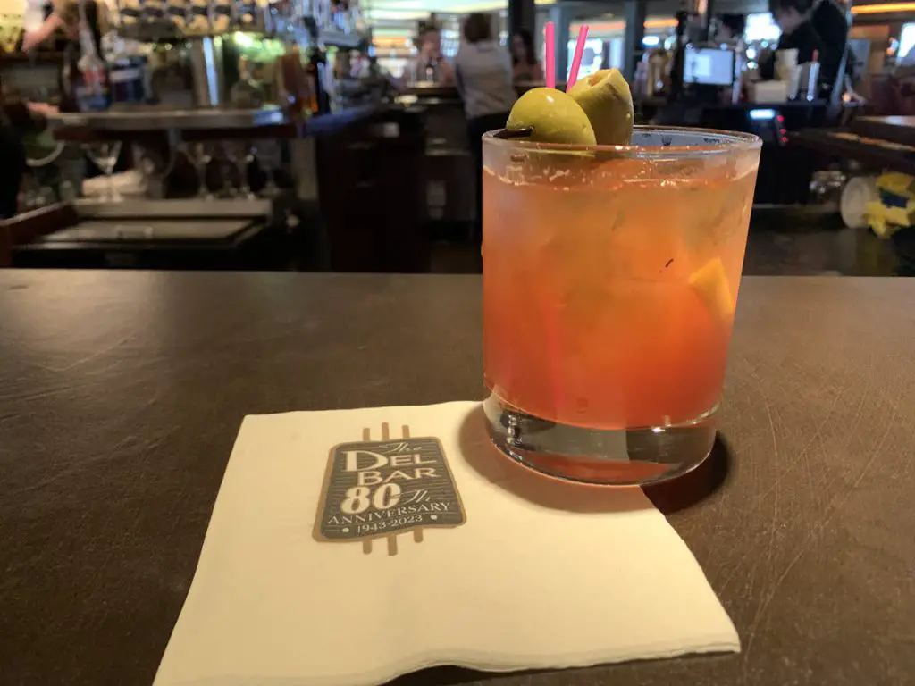 Picture of old fashioned drink with olives on a white napkin that has the Del Bar logo on it.